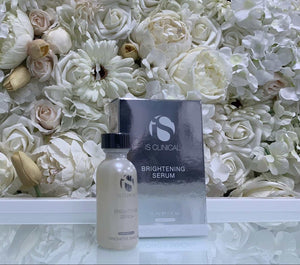 iS Clinical Brightening Serum in a frosted glass bottle with black cap and silver branded box, displayed with white floral background