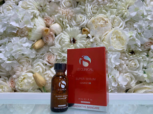 Amber glass bottle with black cap and red box packaging of iS Clinical Super Serum Advance+ multi-tasking vitamin C serum displayed against white and cream rose flowers background.